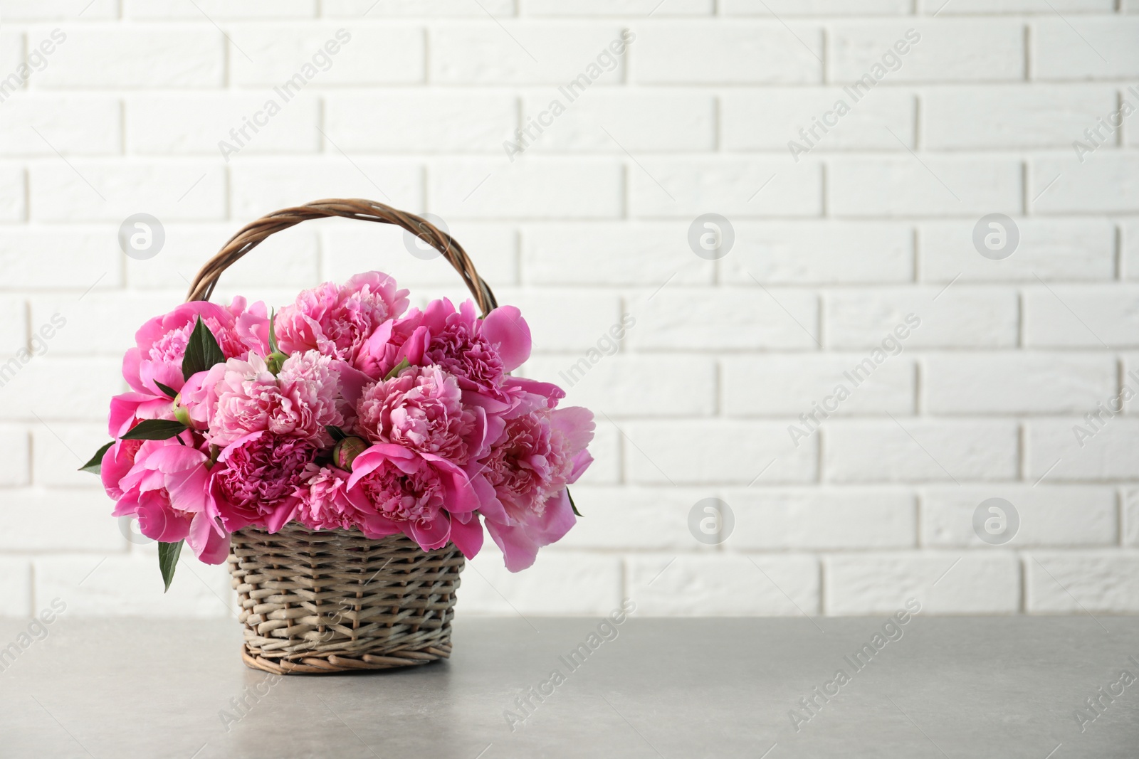 Photo of Wicker basket with fragrant peonies on table against brick wall, space for text. Beautiful spring flowers