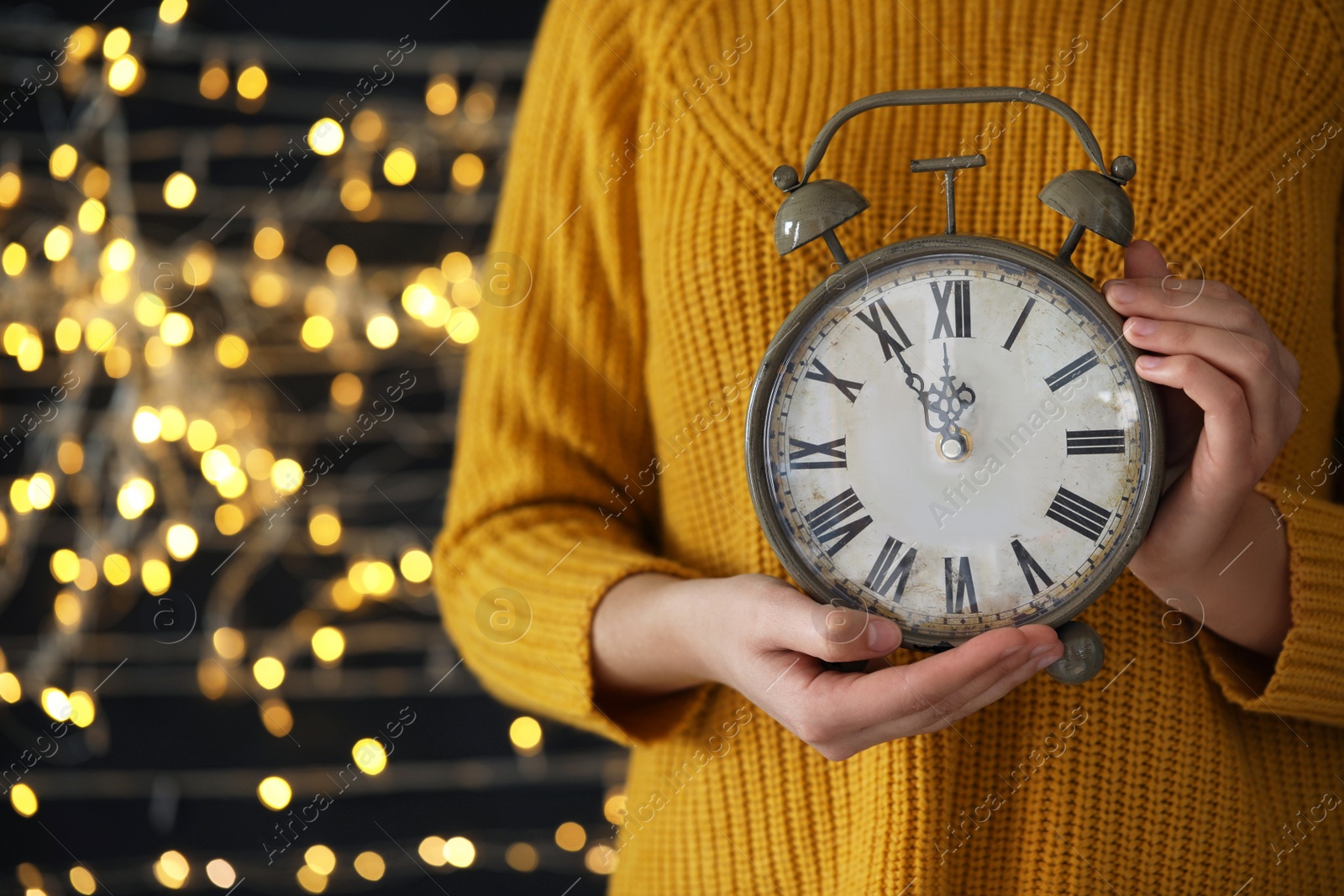 Photo of Woman holding alarm clock against blurred lights, closeup with space for text. New Year countdown