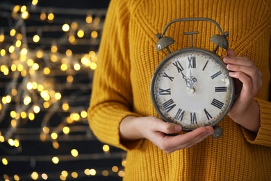 Woman holding alarm clock against blurred lights, closeup with space for text. New Year countdown