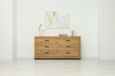 Wooden chest of drawers, lamp and beautiful picture on white wall indoors. Interior design