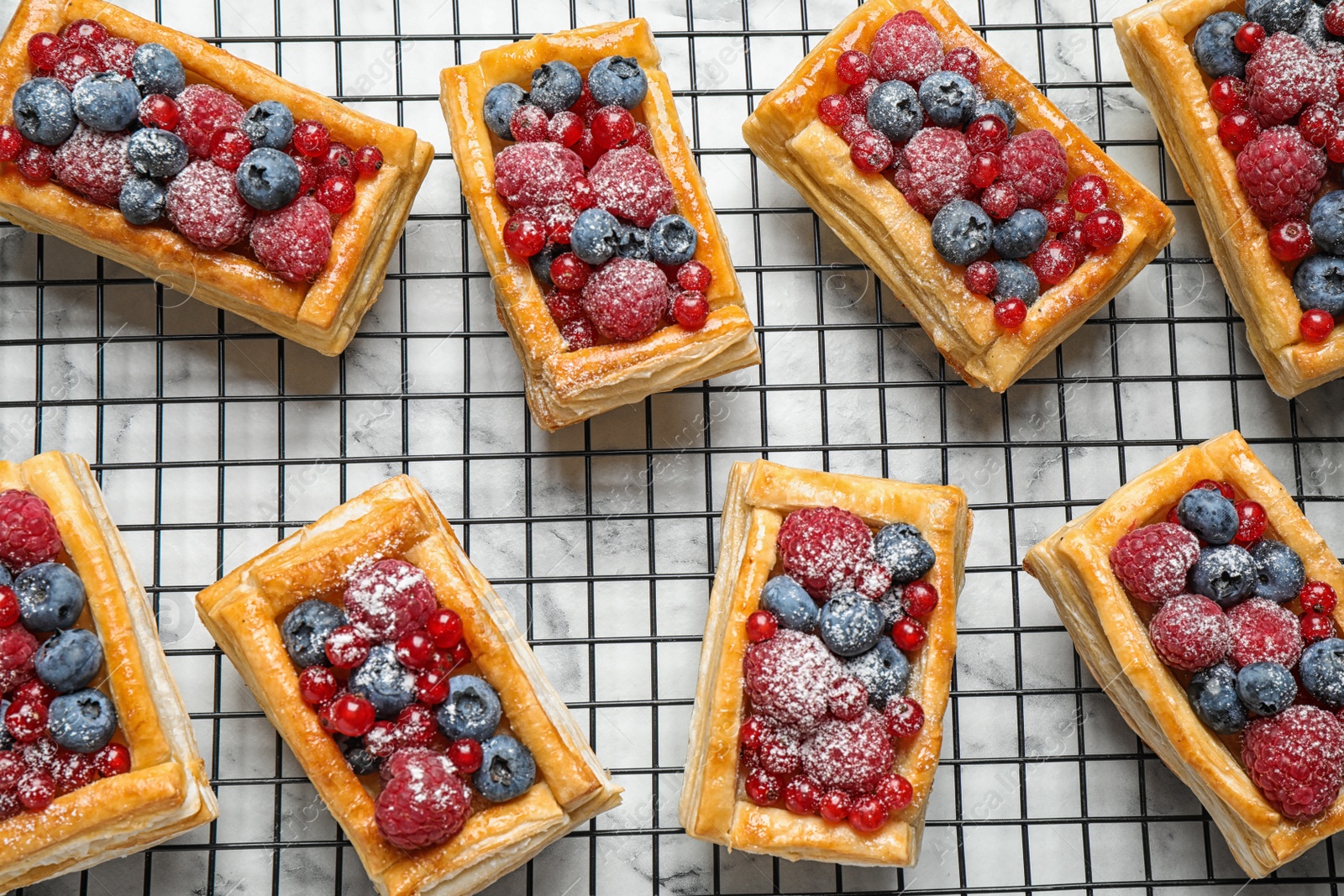 Photo of Cooling rack and fresh delicious puff pastry with sweet berries on white marble table, flat lay