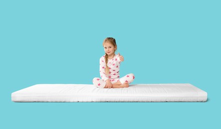 Little girl sitting on mattress and showing thumb up against light blue background