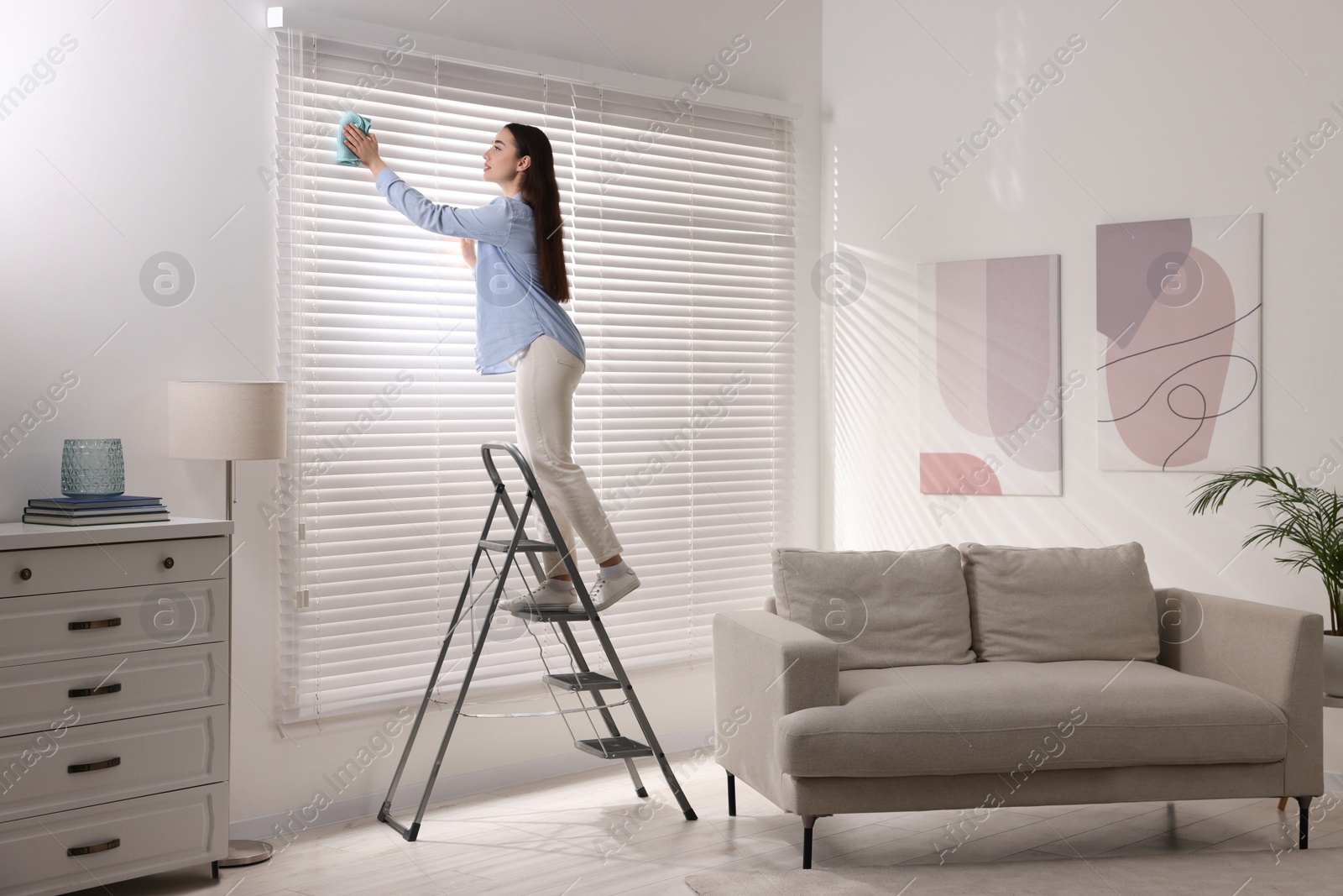 Photo of Woman on metal ladder wiping blinds at home