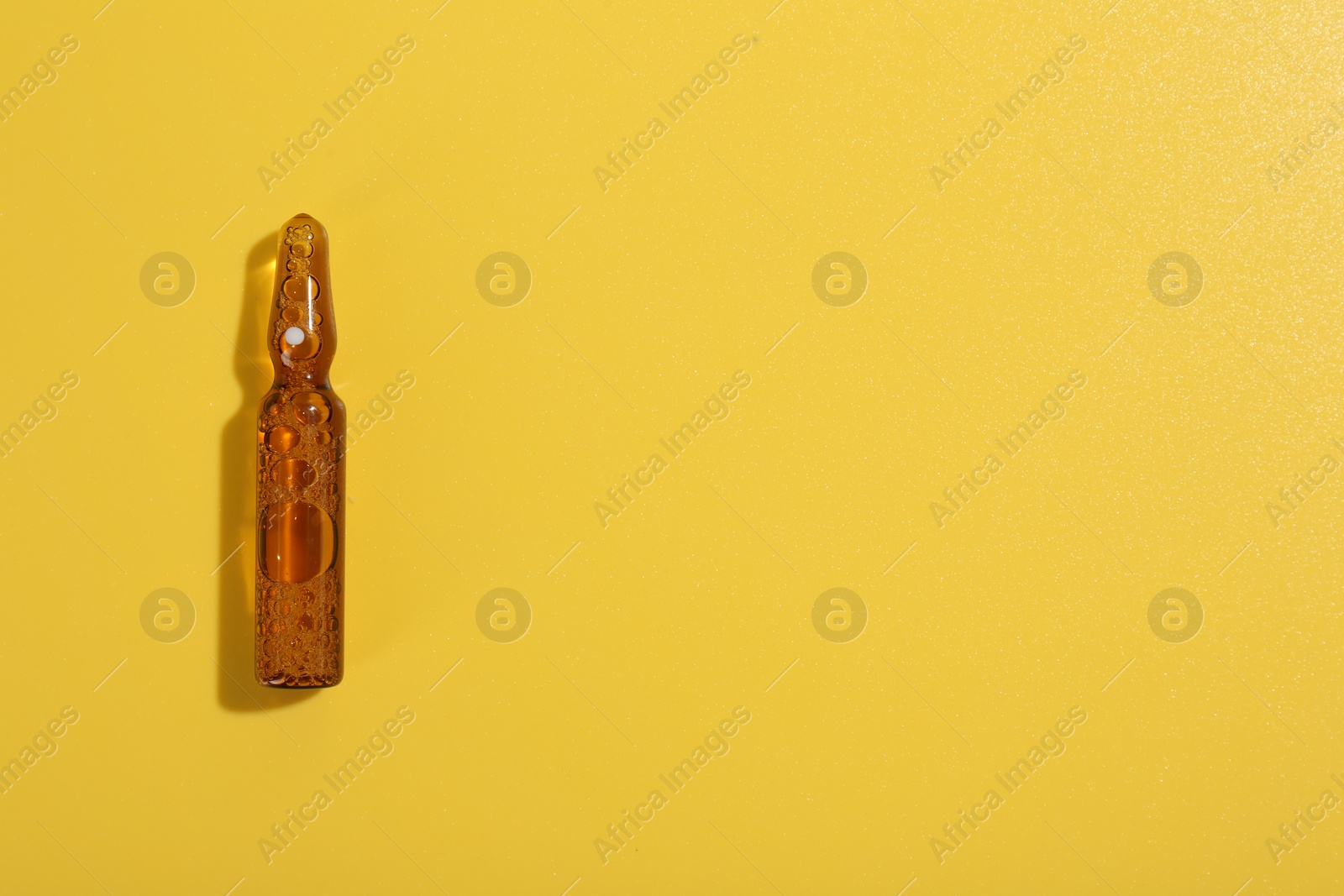 Photo of Glass ampoule with liquid on yellow background, top view. Space for text