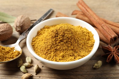 Photo of Dry curry powder in bowl, spoon and other spices on wooden table, closeup