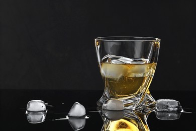 Tasty whiskey and ice cubes in glass on mirror table against black background, closeup. Space for text