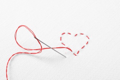 Needle with red thread and stitches in shape of heart on white fabric, top view