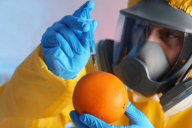 Photo of Scientist in chemical protective suit injecting orange against color background, focus on fruit