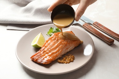 Woman pouring sauce onto tasty cooked salmon on plate, closeup