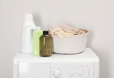 Photo of Baby clothes in wicker basket and laundry detergents on washing machine near light wall