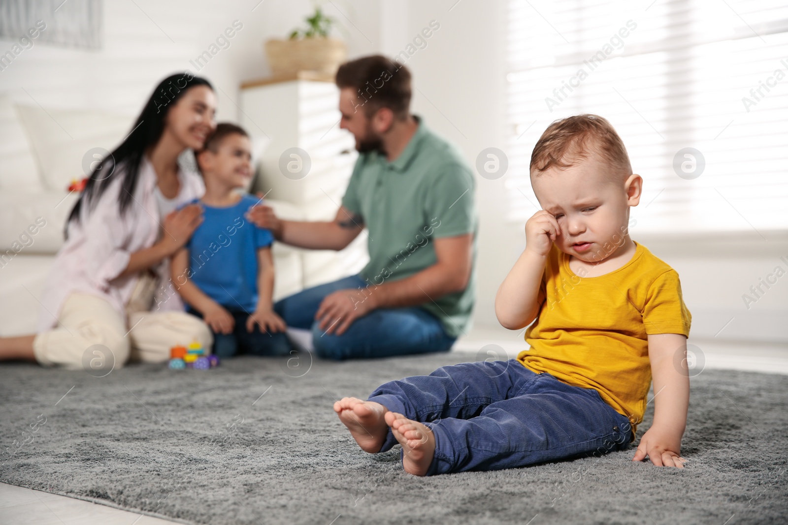 Photo of Unhappy baby sitting alone on floor while parents spending time with his elder brother at home. Jealousy in family