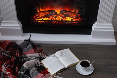 Cup of hot coffee, plaid and book on floor near fireplace, above view. Cozy atmosphere