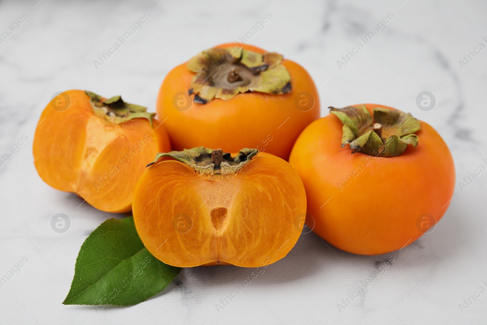 Photo of Whole and cut delicious ripe juicy persimmons with green leaf on white marble table