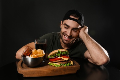 Photo of Young man and tasty burger served on wooden board with French fries against black background