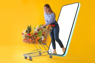 Image of Grocery shopping via internet. Happy woman with shopping cart full of products walking out of huge smartphone on orange background