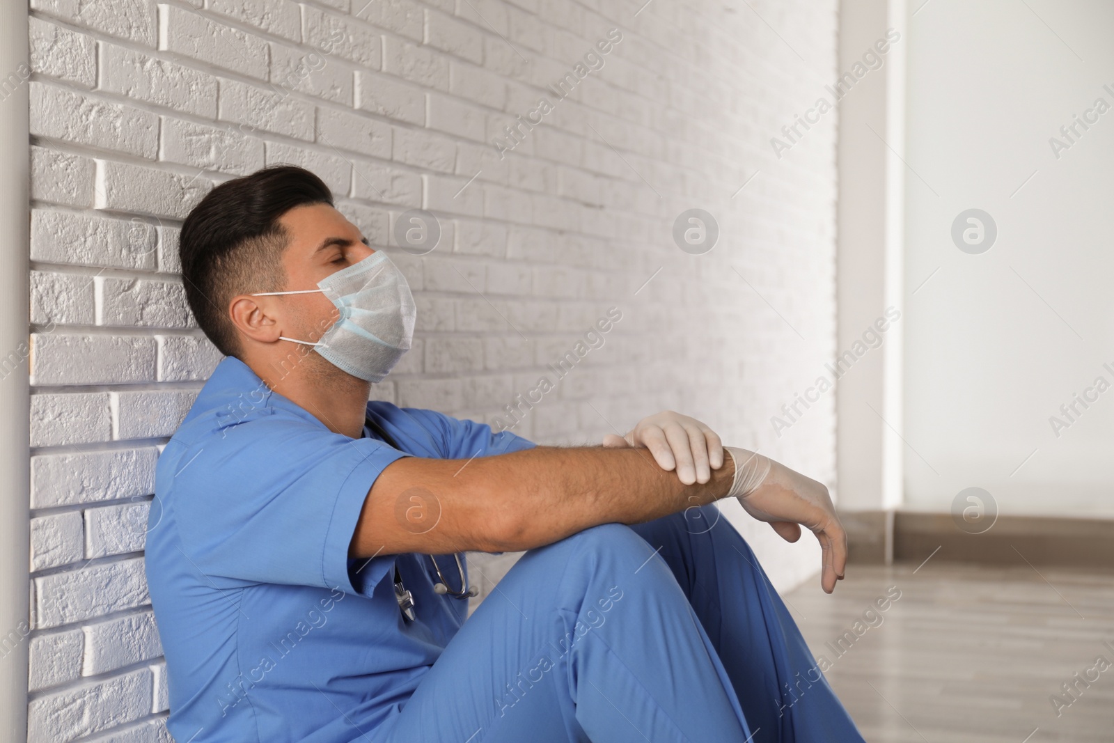 Photo of Sad doctor sitting near white brick wall indoors. Stress of health care workers during coronavirus pandemic