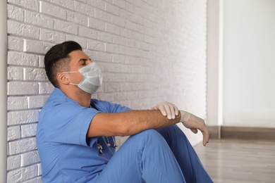 Sad doctor sitting near white brick wall indoors. Stress of health care workers during coronavirus pandemic