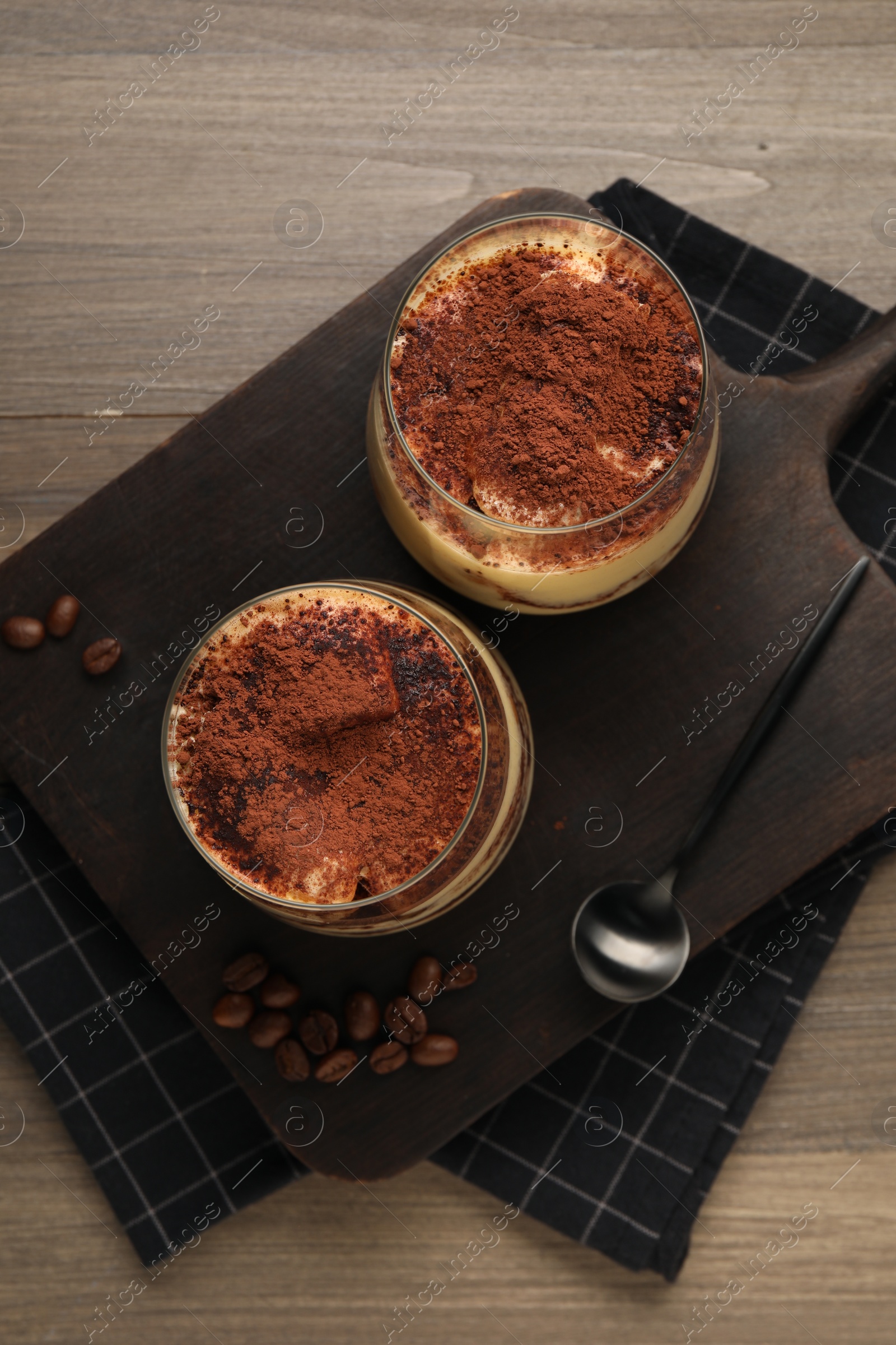 Photo of Delicious tiramisu in glasses, spoon and coffee beans on wooden table, top view