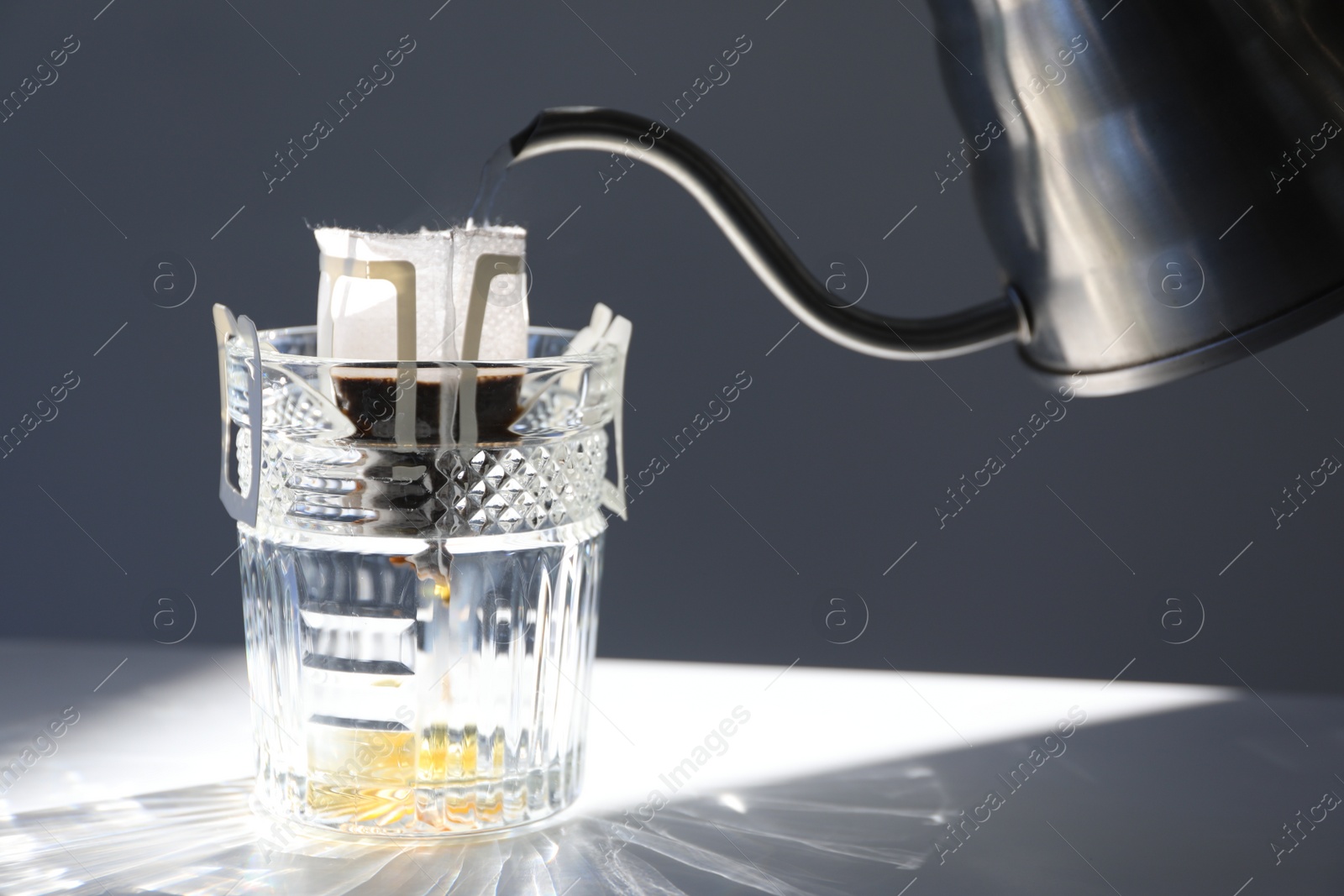 Photo of Pouring hot water into glass with drip coffee bag from kettle on light grey table, closeup