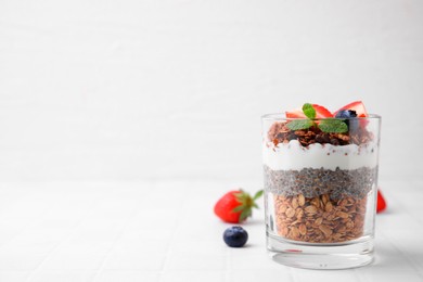 Photo of Tasty granola with berries, yogurt and chia seeds in glass on white tiled table, space for text