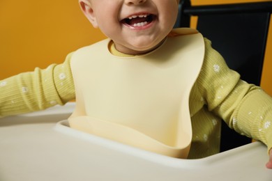 Photo of Cute little baby wearing bib in highchair on yellow background, closeup