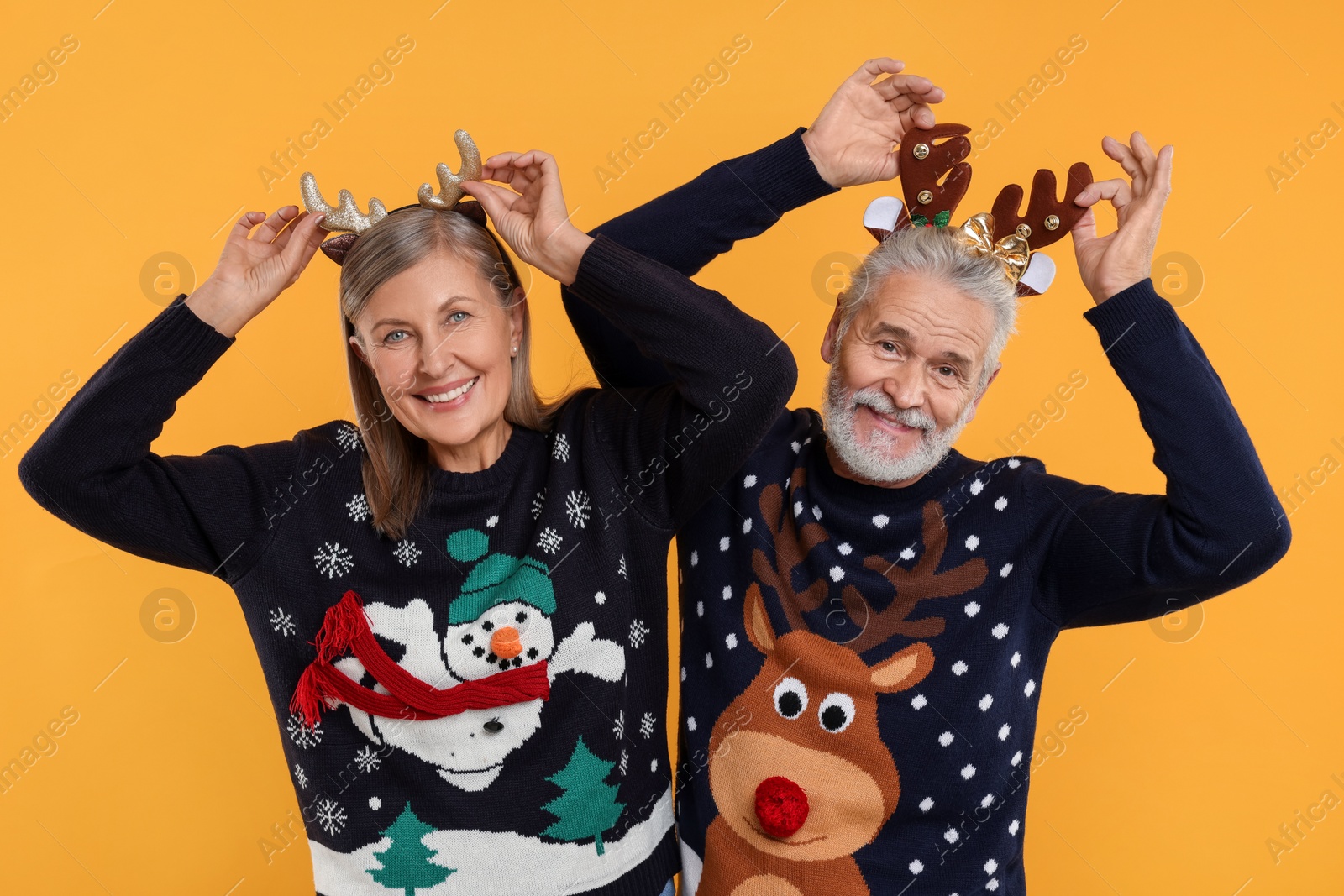 Photo of Senior couple in Christmas sweaters and reindeer headbands on orange background