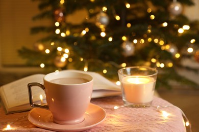 Photo of Cup of delicious cocoa, beautiful candle and open book on pink fabric against blurred Christmas tree with lights