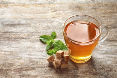 Photo of Cup with hot aromatic mint tea, fresh leaves and sugar cubes on wooden table