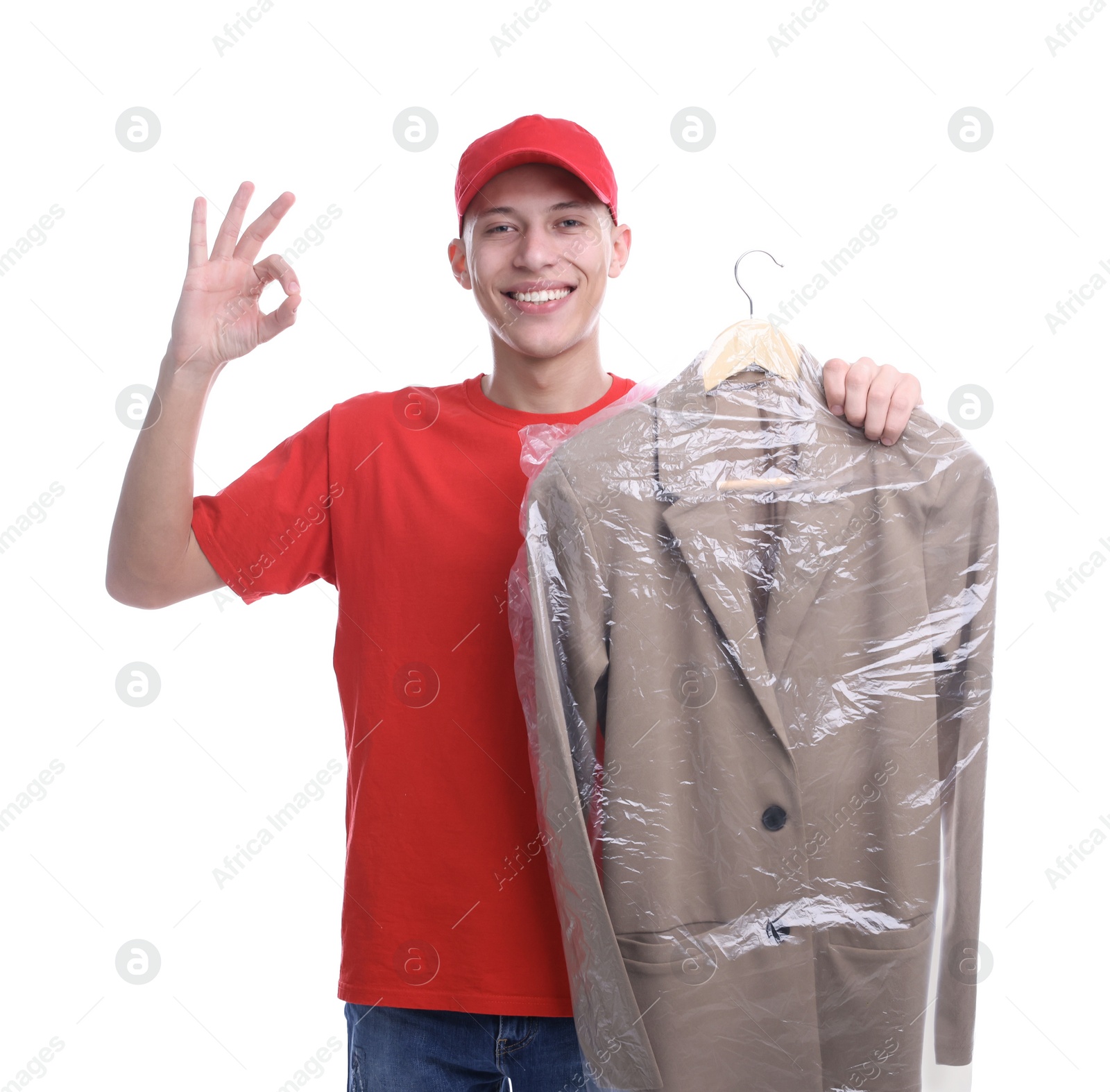 Photo of Dry-cleaning delivery. Happy courier holding jacket in plastic bag and showing OK gesture on white background