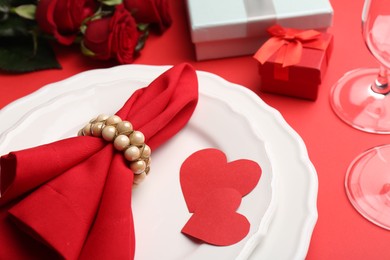 Photo of Plates with napkin, paper hearts, gift boxes and bouquet of roses on red table, closeup. Romantic dinner place setting