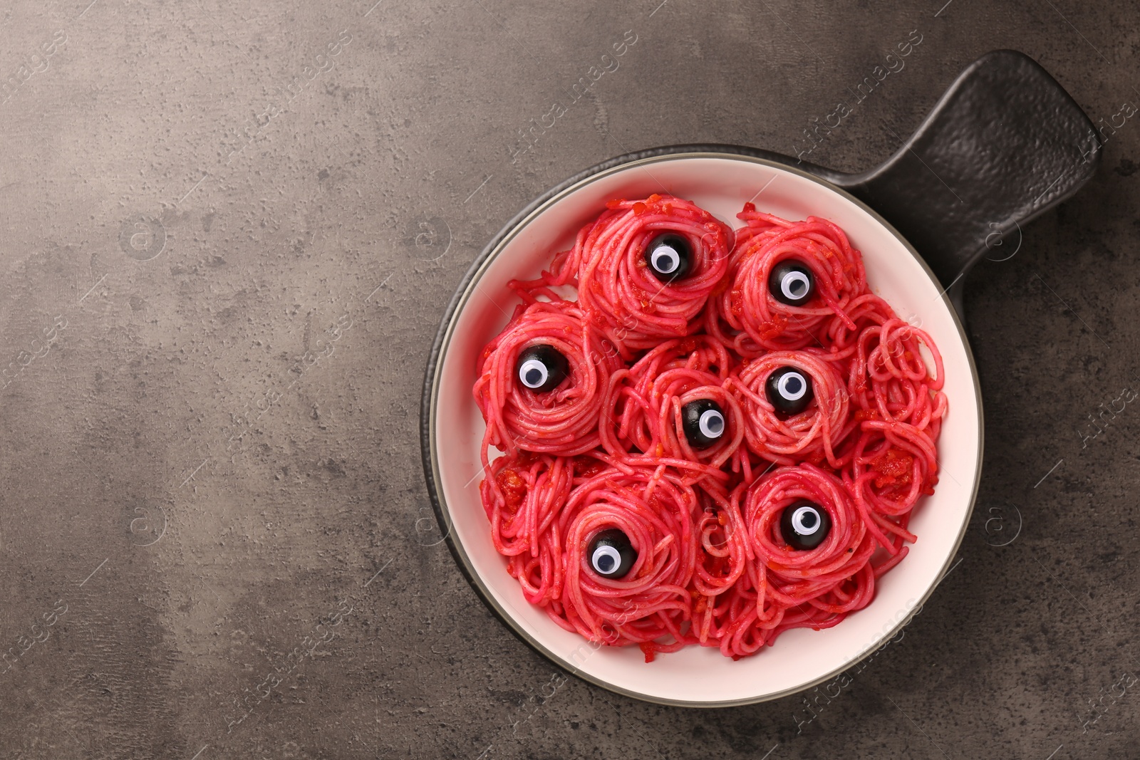 Photo of Red pasta with decorative eyes and olives in bowl on grey textured table, top view with space for text. Halloween food