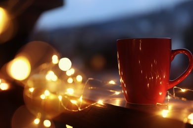 Cup of hot beverage on balcony railing decorated with Christmas lights, space for text. Winter evening