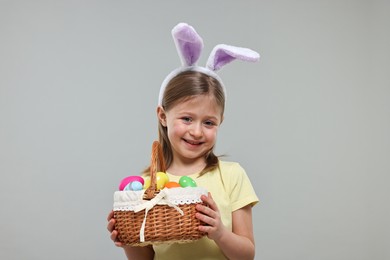 Photo of Easter celebration. Cute girl with bunny ears holding basket of painted eggs on gray background