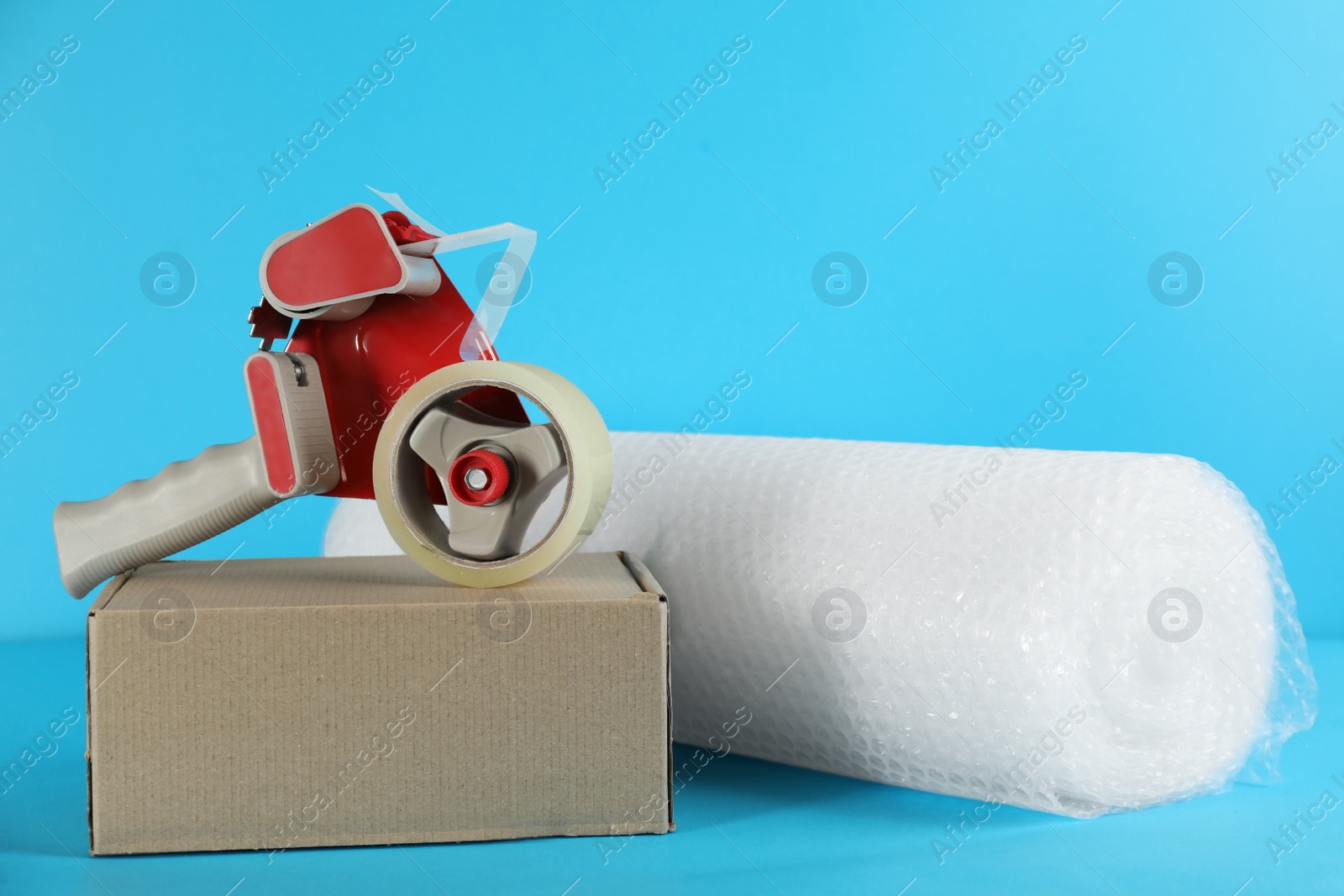 Photo of Bubble wrap roll, tape dispenser and cardboard box on light blue background