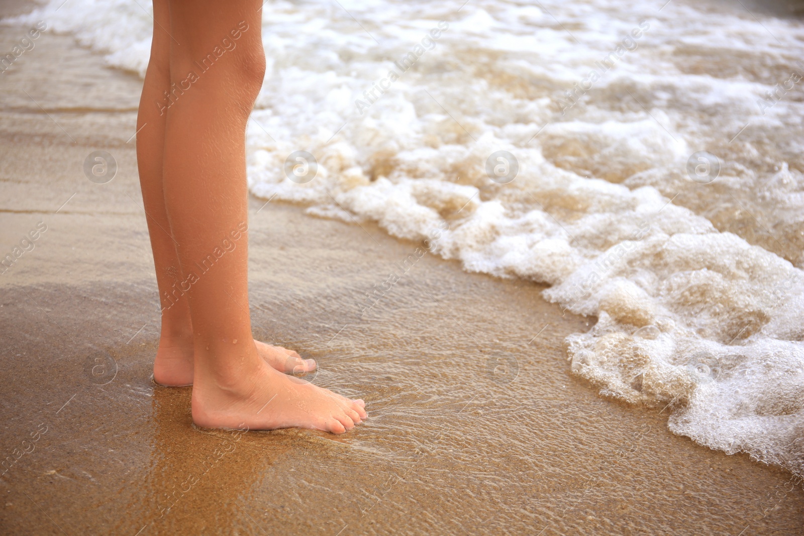 Photo of Little girl standing on sandy beach near sea, closeup. Space for text