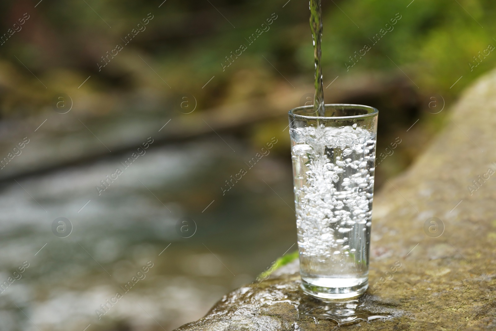 Photo of Fresh water pouring into glass on stone near stream. Space for text