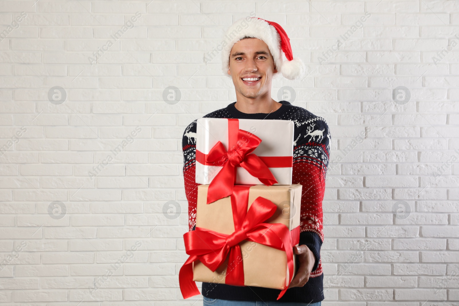 Photo of Happy man in Christmas sweater and Santa hat holding gift boxes near white brick wall