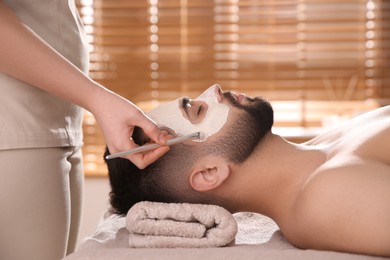 Cosmetologist applying mask on man's face in spa salon