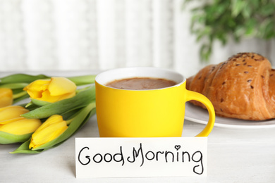 Photo of Croissant, coffee, beautiful flowers and card with words GOOD MORNING on white wooden table indoors