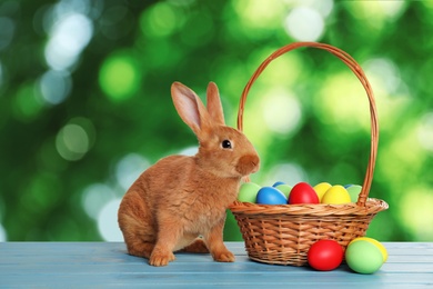 Adorable bunny and wicker basket with Easter eggs on wooden surface outdoors