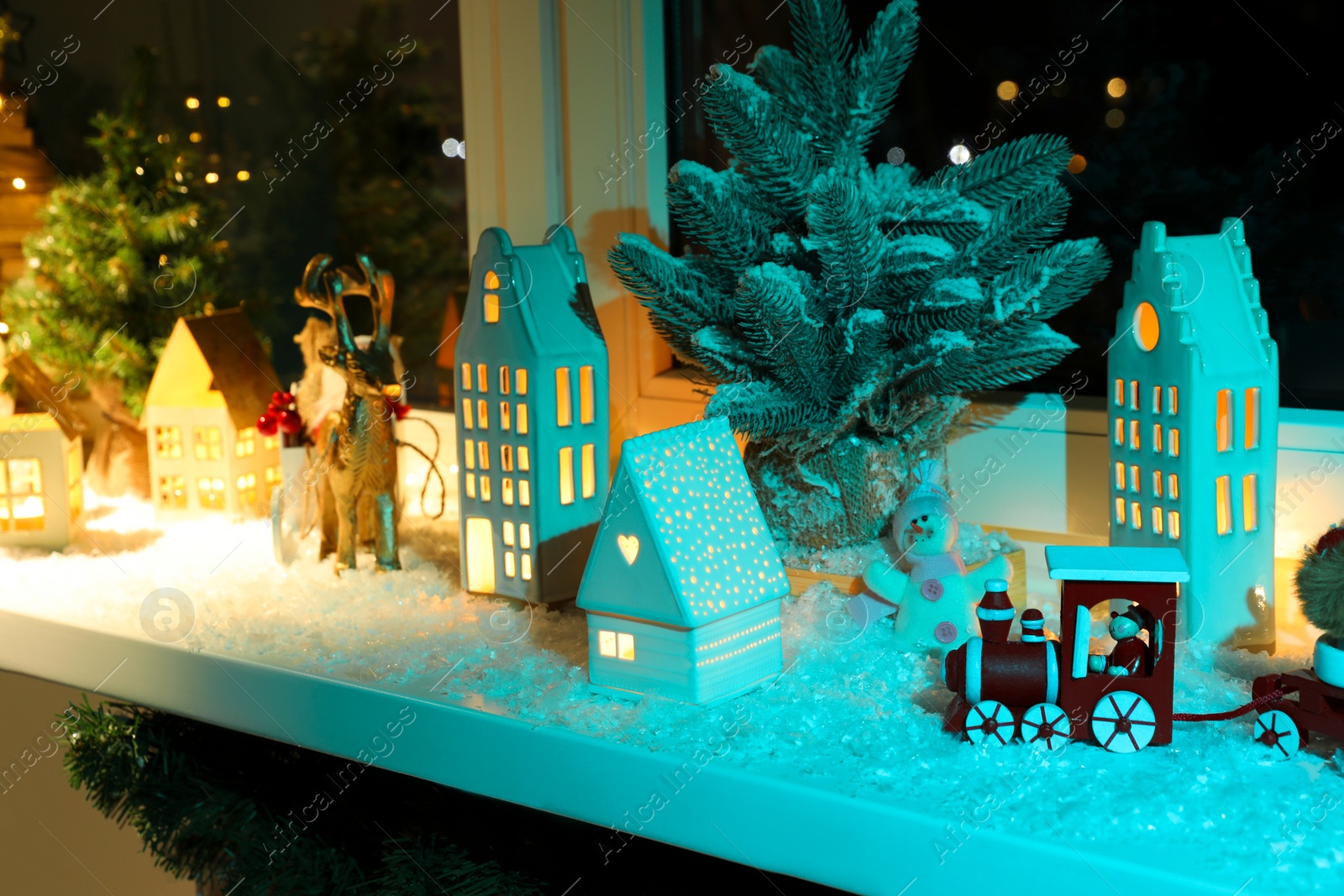 Photo of Christmas atmosphere. Beautiful glowing houses, fir trees, artificial snow and toys on window sill indoors