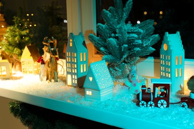 Christmas atmosphere. Beautiful glowing houses, fir trees, artificial snow and toys on window sill indoors