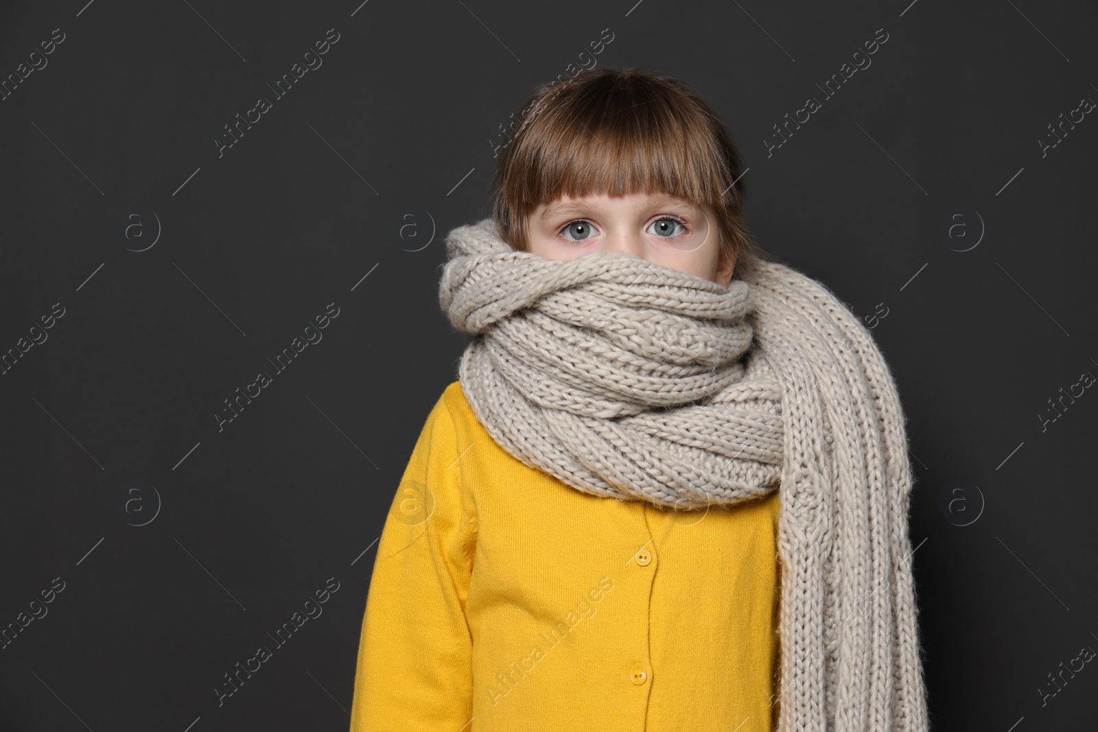 Photo of Funny little girl wrapped in scarf against dark  background, space for text. Cough precaution