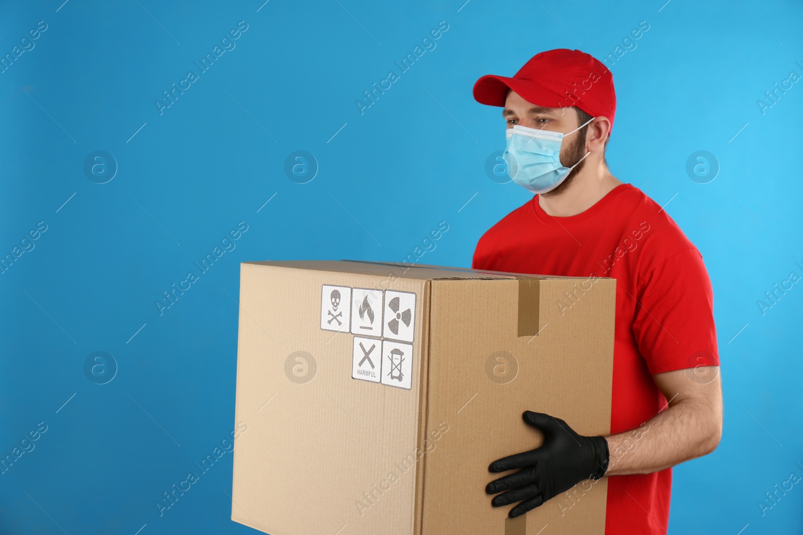 Photo of Courier in mask holding cardboard box with different packaging symbols on blue background. Parcel delivery