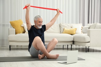 Photo of Senior man doing exercise with fitness elastic band near laptop on mat at home