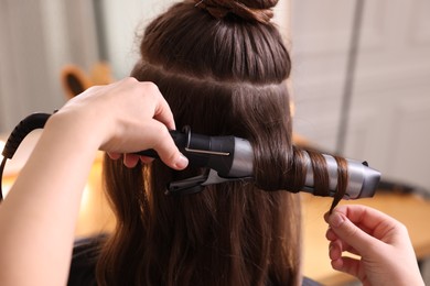 Photo of Hairdresser using curling hair iron while working with woman in salon, closeup