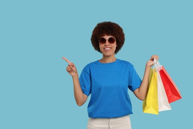 Happy young woman with shopping bags pointing at something on light blue background. Space for text