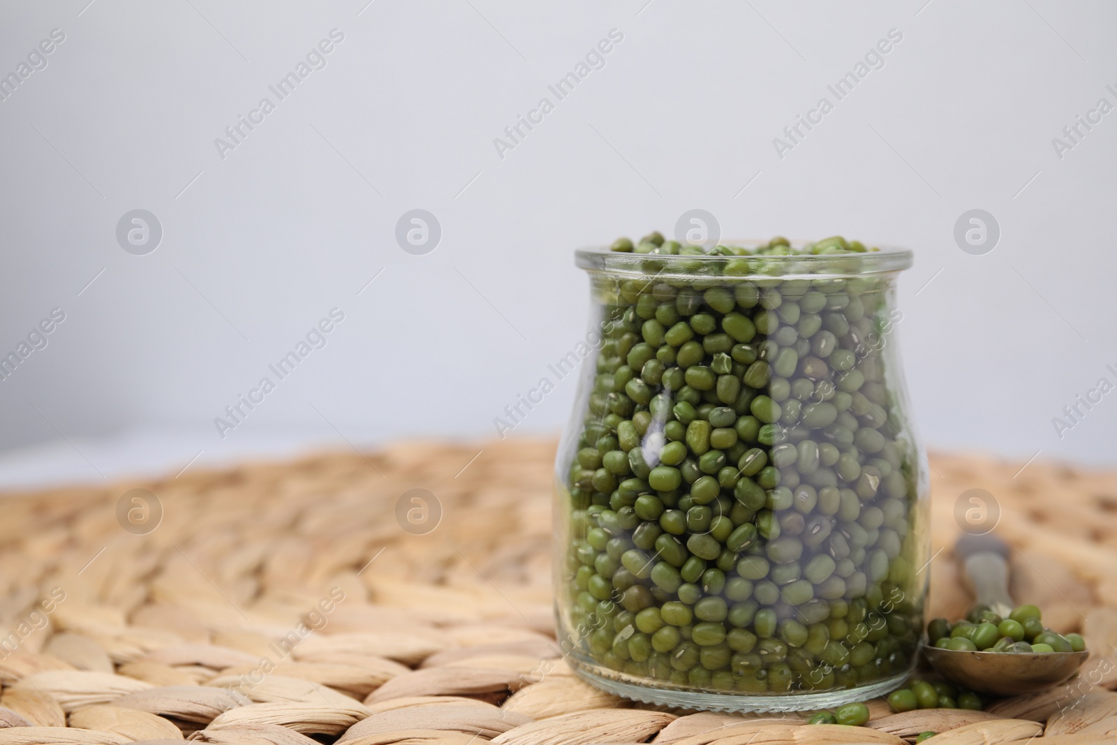 Photo of Glass jar of mung beans with spoon on wicker mat against white background, closeup. Space for text