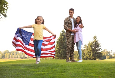 Little girl running with American flag, her father in military uniform and mother at sunny park