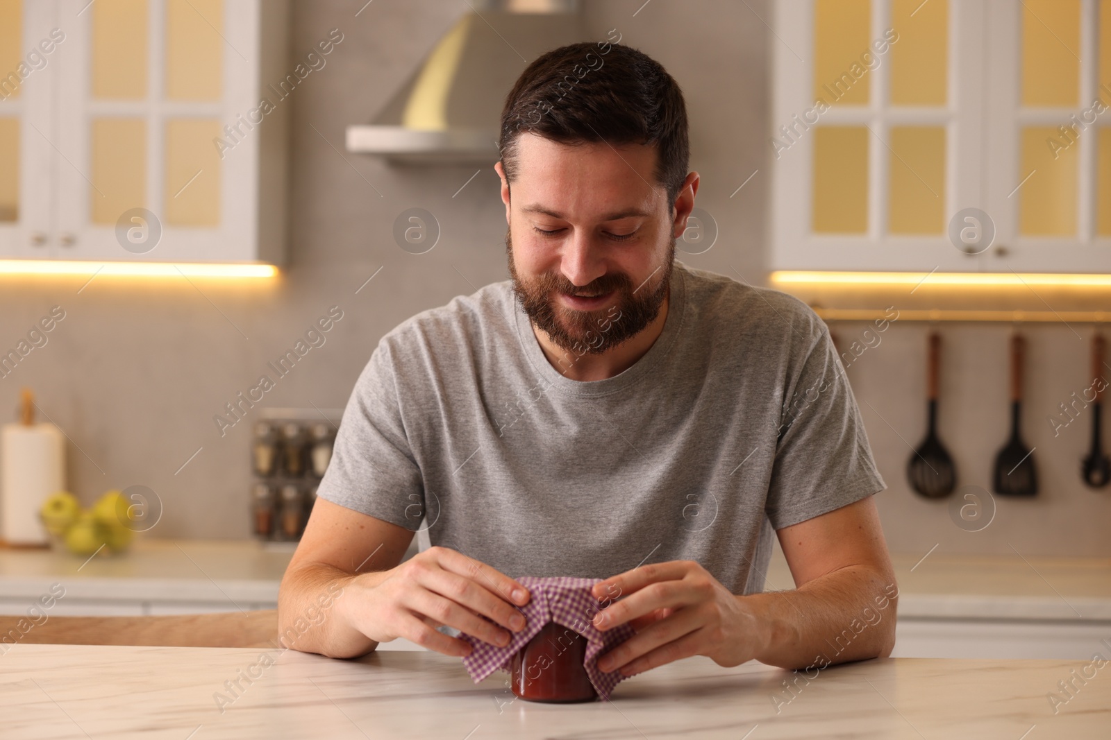 Photo of Man packing jar of jam into beeswax food wrap at table in kitchen